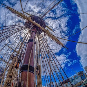 May include: A close-up view of the mast and rigging of a tall ship, with a blue sky and white clouds in the background. The ship's mast is made of wood and is covered in ropes. The ropes are attached to the mast and to the sails.