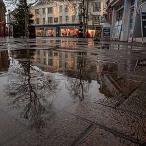 May include: A wet, brick-paved street with a reflection of a building with Christmas lights in a puddle. The reflection is distorted by the water.