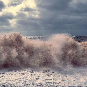 Könnte beinhalten: Eine große Welle bricht an der Küste und erzeugt einen Sprühnebel aus weißem Schaum und eine Wolke aus braunen Wassertröpfchen. Der Himmel ist mit dunklen Wolken bedeckt, aber ein Sonnenstrahl blitzt durch.