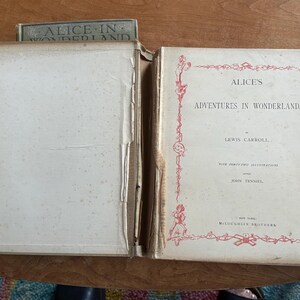 May include: An open vintage book titled "Alice's Adventures in Wonderland" by Lewis Carroll. The cover is off-white with red decorative borders and text. The book is displayed open, revealing aged, off-white pages.