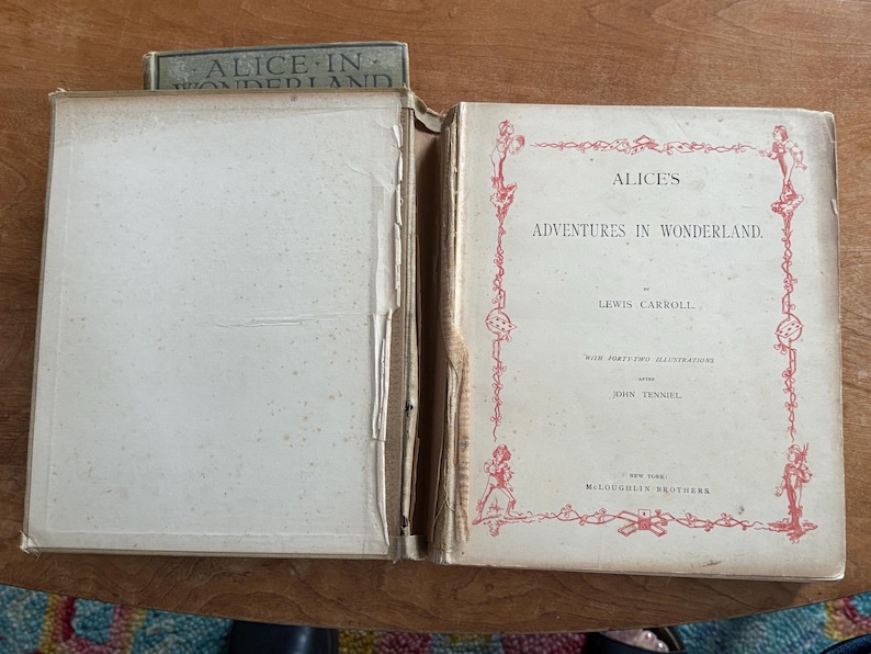 May include: An open vintage book titled "Alice's Adventures in Wonderland" by Lewis Carroll. The cover is off-white with red decorative borders and text. The book is displayed open, revealing aged, off-white pages.
