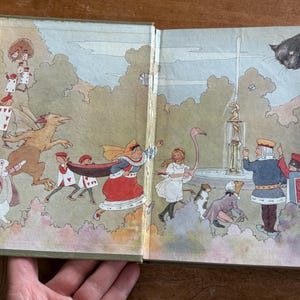 May include: Open book illustration featuring characters from Alice in Wonderland. The artwork depicts the Queen of Hearts, Alice, the White Rabbit, and playing card soldiers. A black cat floats in the sky above a fountain.