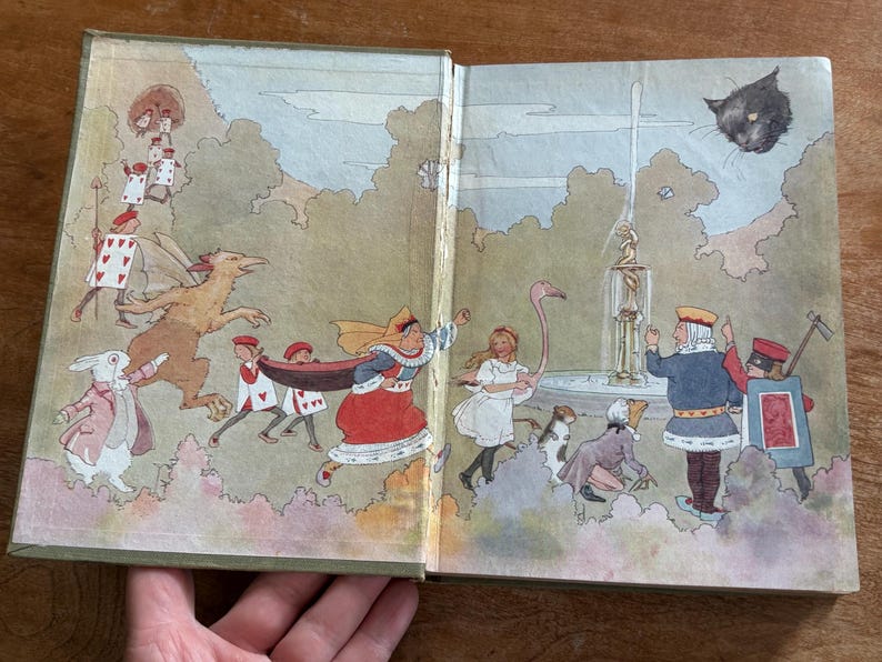 May include: Open book illustration featuring characters from Alice in Wonderland. The artwork depicts the Queen of Hearts, Alice, the White Rabbit, and playing card soldiers. A black cat floats in the sky above a fountain.