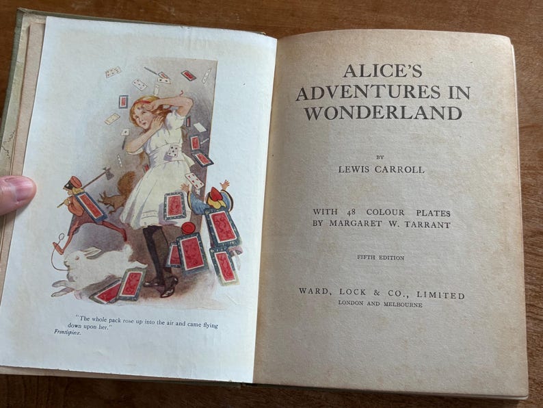 May include: Open book with illustrations from "Alice's Adventures in Wonderland." The left page shows Alice in a white dress, surrounded by playing cards. The right page displays the title and author, Lewis Carroll.