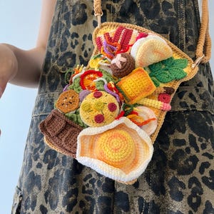 Puede incluir: Un bolso bandolera de ganchillo hecho a mano con forma de rebanada de pan tostado, adornado con varios alimentos. El bolso tiene una correa marrón y está lleno de comida de ganchillo colorida y detallada, incluyendo un huevo frito, pizza y verduras.