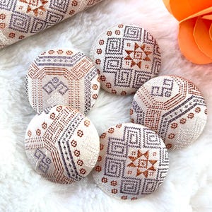 May include: Five round, white fabric buttons with a geometric pattern in shades of orange, purple, and white. The buttons are arranged in a circle on a white fuzzy surface.