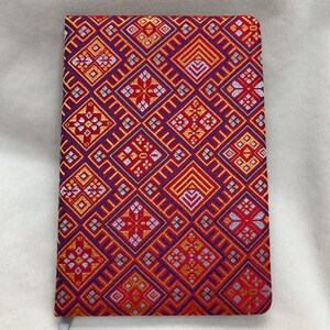 May include: A red, blue, and gold patterned notebook with a silver bookmark. The cover is made of a woven fabric with a geometric design.