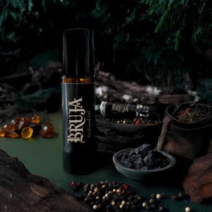 Puede incluir: Un frasco negro de "BRUJA Perfumed Oil" en formato roll-on sobre una superficie verde con granos de pimienta negra y blanca, piedras de ámbar y un pequeño cuenco negro lleno de piedras negras.