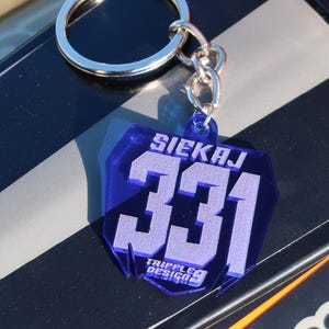 Puede incluir: Un llavero de plástico azul con el nombre "Siekaj" y el número "331" en letras blancas. El llavero también tiene el texto "Triple9 Design" en letras blancas.