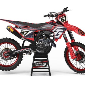 Op de afbeelding: Rode en zwarte motorcrossmotor met nummer 67 op de voor- en zijpanelen. De motor heeft zwarte banden, een zwart zadel en een zwarte motor. De velgen zijn rood. De motor staat op een zwarte standaard.