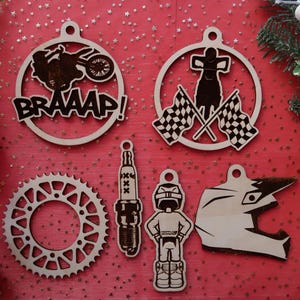 May include: A collection of wooden motocross-themed decorations on a red surface. The ornaments feature a motorcycle with the word "BRAAAP!", a rider with chequered flags, a sprocket, a spark plug, a rider, and a helmet. Designed for hanging.