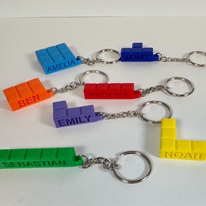 May include: Six colourful personalised keychains made of plastic building blocks. The keychains feature the names Amelia, Ben, Emily, James, Noah, and Phoenix.