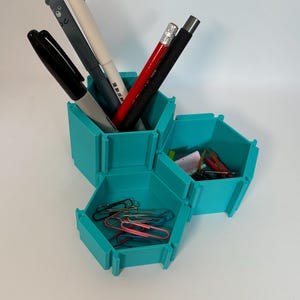 May include: Teal hexagonal desk organiser with five compartments. The organiser holds pens, pencils, and paper clips.