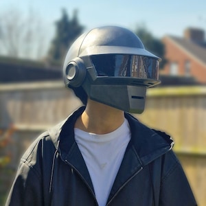 Puede incluir: Un casco de estilo futurista gris oscuro, con visera y detalles laterales circulares. El casco se lleva con una chaqueta negra y una camiseta blanca. El fondo es una escena al aire libre soleada.