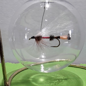 May include: A decorative clear glass orb with a fishing fly suspended inside. The fly has white, brown, and red accents. The orb is held by a gold-colored metal stand, creating a unique display piece.