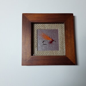 May include: A framed fly fishing lure. The square frame is made of dark brown wood. The lure has orange and red feathers, a silver body, and a black hook. The background is a burlap-like material.