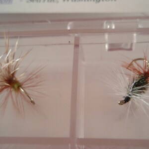 May include: Two artificial fishing flies displayed in a clear plastic container. The flies have intricate designs with brown, white, and orange fibers, and small metal hooks. The flies are designed to mimic insects and attract fish.