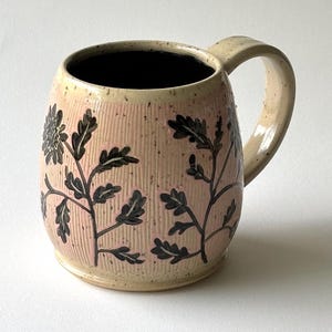 Könnte beinhalten: Eine handgefertigte Keramiktasse mit hellrosa Hintergrund und schwarzem Blumendesign. Die Tasse hat einen beigen Griff und Rand, und der Innenraum ist schwarz. Die Tasse ist zylindrisch mit einer strukturierten Oberfläche.
