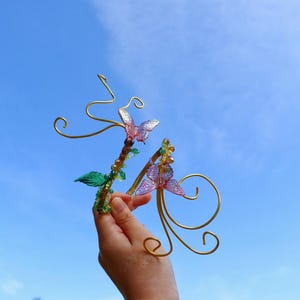 Peut inclure: Deux accessoires décoratifs pour cheveux en forme de papillon. Chacun présente une monture en fil d'or avec des papillons et des feuilles. Les papillons sont roses et violets. Le fond est un ciel bleu clair.