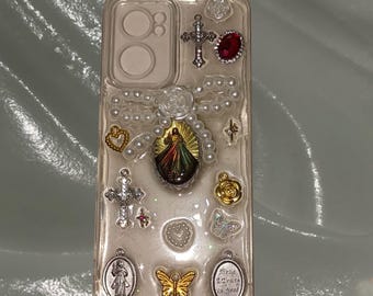 May include: A clear phone case with a pearl and gold chain design. The case features a variety of charms, including a cross, a heart, a butterfly, and a religious image. The case is decorated with glitter and has a red gem.