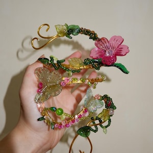 May include: A floral-themed hair accessory with a gold wire frame. The accessory features pink flowers, green leaves, and clear butterfly accents, adorned with colorful beads. The design is intricate and delicate, suitable for special occasions.