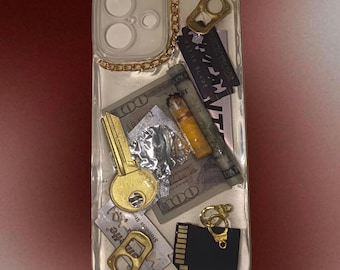 May include: Clear phone case with embedded objects including a gold key, a hundred-dollar bill, a razor blade, a cigarette, and a gold chain. The case also features a gold bottle opener and a black SD card.