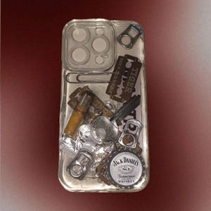 May include: Clear phone case filled with various small objects, including a Jack Daniel's bottle cap, razor blades, and a cigarette. The case has a clear, transparent design, showcasing the items inside. The case has a camera cutout.