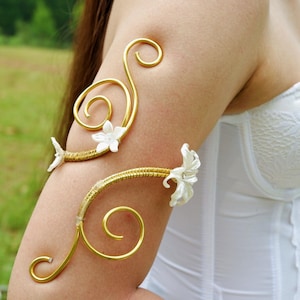 May include: Gold-toned arm cuffs with a floral design. The cuffs feature a swirling, openwork design with white flower accents. The arm cuffs are worn on the upper arm and are set against a background of green foliage.