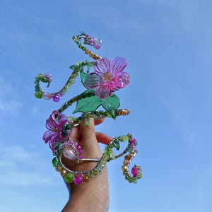 May include: A decorative floral hair accessory featuring pink and purple flowers, green leaves, and colourful beads. The accessory is held against a clear blue sky, highlighting its intricate design and vibrant colours.