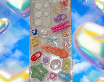 May include: Clear phone case with embedded colorful charms, including a pink clothespin, orange barrette, and a green star. Other charms include a butterfly, bow, and gummy bears. The case is decorated with glitter.