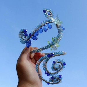 May include: A handcrafted arm cuff with a spiral design. The cuff is made of silver-colored wire and adorned with blue and clear beads of various shapes and sizes. The beads are arranged to create a floral and droplet design.