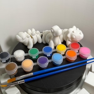 May include: A craft kit featuring white dinosaur figurines, small paint pots in various colors, and two blue-handled paintbrushes. The paint pots are arranged in a row, ready for use. White foam blocks are in the foreground.