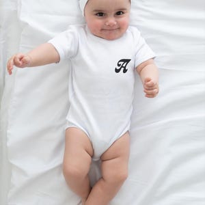 May include: A baby wearing a white onesie with a black letter 'A' on the front. The baby is wearing a white headband with a bow. The baby is lying on a white sheet.