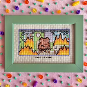 May include: A cross-stitch embroidery in a sage green frame featuring a brown dog sitting in a room filled with orange and yellow flames. The text "THIS IS FINE" is stitched in black thread below the dog, set against a light fabric background.