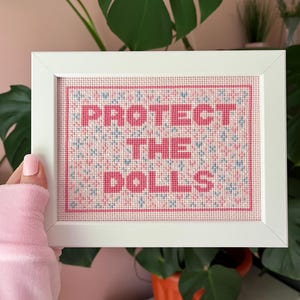 Può includere: Un'opera d'arte a punto croce incorniciata con le parole "PROTECT THE DOLLS" in rosa. Lo sfondo presenta un motivo di punti croce rosa, blu e bianchi. La cornice è bianca e l'opera è esposta su uno sfondo di piante verdi.