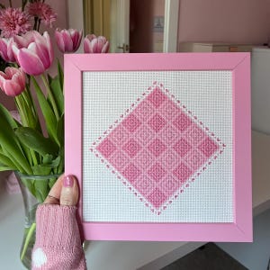 May include: A pink framed cross-stitch featuring a diamond-shaped plaid pattern in varying shades of pink. The artwork is displayed against a white background and framed in a solid pink. The image is set against a background of pink tulips in a glass vase.