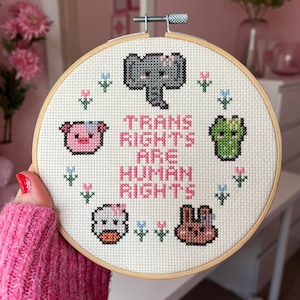 以下が含まれることがあります： ピンク色の「TRANS RIGHTS ARE HUMAN RIGHTS」の文字が入ったクロスステッチ刺繍枠。象、豚、カエル、アヒル、ウサギのピクセル化された動物のキャラクターと花のアクセントがデザインされています。
