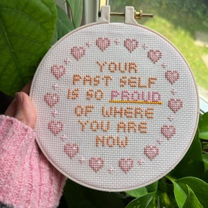 K&ouml;nnte beinhalten: Ein Kreuzstich-Stickrahmen mit dem Text "YOUR PAST SELF IS SO PROUD OF WHERE YOU ARE NOW" in orangefarbenem und rosa Garn. Das Design umfasst rosa Herzen um den Rand. Der Rahmen wird vor einem Fenster gehalten.