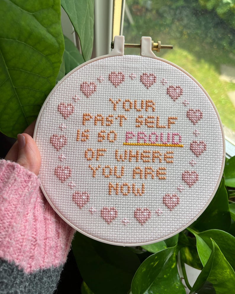 K&ouml;nnte beinhalten: Ein Kreuzstich-Stickrahmen mit dem Text "YOUR PAST SELF IS SO PROUD OF WHERE YOU ARE NOW" in orangefarbenem und rosa Garn. Das Design umfasst rosa Herzen um den Rand. Der Rahmen wird vor einem Fenster gehalten.