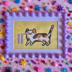 Könnte beinhalten: Eine Kreuzstichstickerei einer Glückskatze, die eine Trans-Pride-Flagge im Maul hält, vor einem gelben Stoffhintergrund. Das Werk befindet sich in einem hellvioletten Rahmen mit Perlenrand, umgeben von verstreuten, bunten Kunststoffperlen.