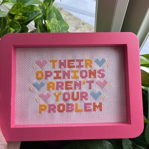 Puede incluir: Un bordado de punto de cruz enmarcado en rosa con el texto "THEIR OPINIONS AREN'T YOUR PROBLEM" en varios colores. La pieza presenta detalles de corazones y estrellas. El marco es de color rosa brillante.