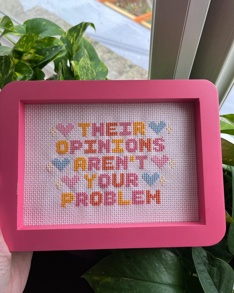 Puede incluir: Un bordado de punto de cruz enmarcado en rosa con el texto "THEIR OPINIONS AREN'T YOUR PROBLEM" en varios colores. La pieza presenta detalles de corazones y estrellas. El marco es de color rosa brillante.