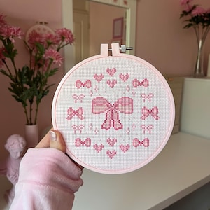 Op de afbeelding: Een roze borduurring met een kruissteekontwerp van een grote strik omringd door kleinere strikken en harten. Het ontwerp is op witte stof, omlijst door een roze ring. Het thema is vrouwelijk en decoratief.