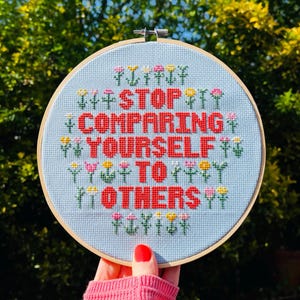 Pode incluir: Um aro de bordado azul com a frase "STOP COMPARING YOURSELF TO OTHERS" em letras vermelhas rodeado de pequenas flores coloridas.