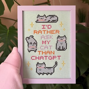Pode incluir: Uma bordada em ponto cruz emoldurada, com uma moldura rosa claro, apresentando quatro gatos cinzentos e o texto "I'd rather ask my cat than ChatGPT". O fundo &eacute; verde claro.