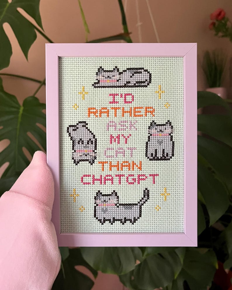Pode incluir: Uma bordada em ponto cruz emoldurada, com uma moldura rosa claro, apresentando quatro gatos cinzentos e o texto "I'd rather ask my cat than ChatGPT". O fundo &eacute; verde claro.