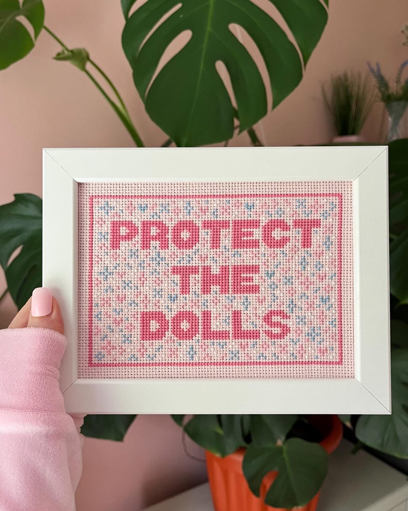 Puede incluir: Una obra de arte en punto de cruz enmarcada con las palabras "PROTECT THE DOLLS" en rosa. El fondo presenta un patr&oacute;n de puntadas de cruz rosas, azules y blancas. El marco es blanco y la obra se exhibe sobre un fondo de plantas verdes.