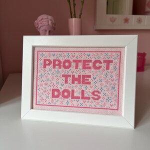 Puede incluir: Una obra de arte de punto de cruz enmarcada con las palabras "PROTECT THE DOLLS" en letras rosas. El fondo es una mezcla de punto de cruz rosa y azul. El marco es blanco y la obra se exhibe sobre una superficie blanca.