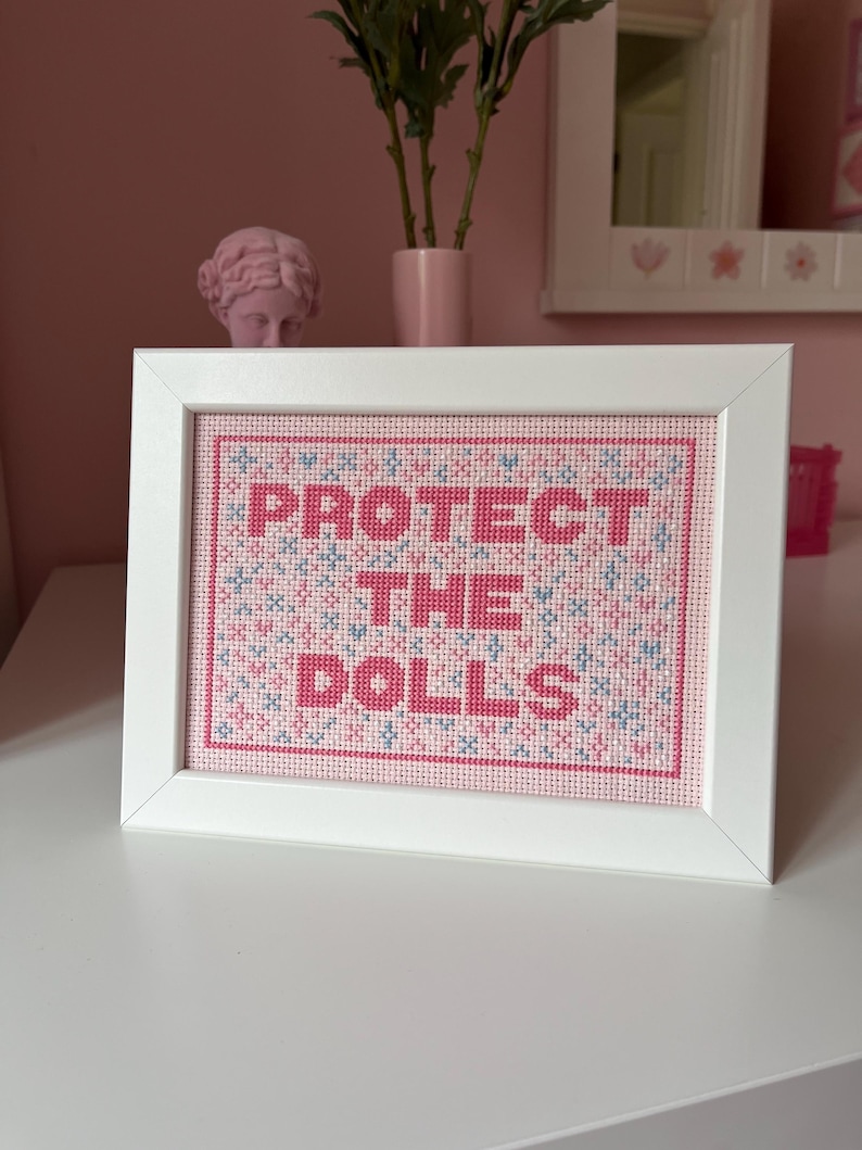 Puede incluir: Una obra de arte de punto de cruz enmarcada con las palabras "PROTECT THE DOLLS" en letras rosas. El fondo es una mezcla de punto de cruz rosa y azul. El marco es blanco y la obra se exhibe sobre una superficie blanca.