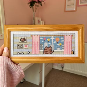 May include: Cross-stitch artwork in a wooden frame. The design features a cat looking out a window with pink curtains, hot air balloons, and a cityscape. Additional elements include a cat silhouette, hearts, and the text "MEOW MEOW".